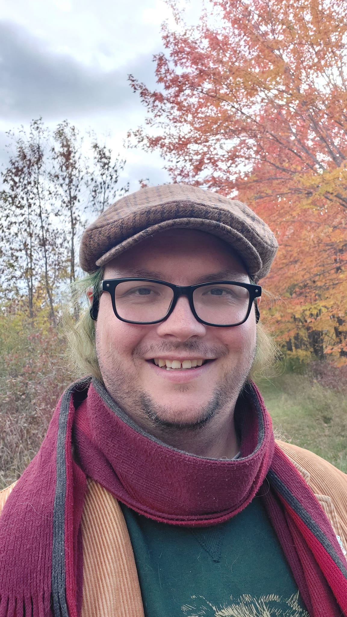 A man with long green hair wearing a green sweater, brown corderoy jacket and red scarf stands in front of a tree who's leaves have turned all colours, red through yellow, Photo 1